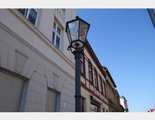 Straßenszene mit einer historischen Laterne im Vordergrund, flankiert von hellen Gebäuden mit dekorativen Fassadenelementen und Ziegelmustern; leichter blauer Himmel im Hintergrund.