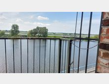 Blick durch ein Geländer auf einen breiten Fluss mit ruhiger Wasseroberfläche, umgeben von einer grünen Uferlandschaft und einigen Bäumen. Rechts ist eine Backsteinwand zu sehen, im Hintergrund befinden sich ein modernes Gebäude und eine kleine Anlegestelle mit Booten.