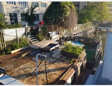 Ein altes Hausboot am Ufer eines Kanals, mit Pflanzenkästen auf dem Dach und einer Vielzahl von Gegenständen, darunter ein Trampolin, auf dem Deck. Im Hintergrund sind ein Zaun, eine Treppe, ein begrünter Gehweg und Gebäude mit Fenstern sichtbar.