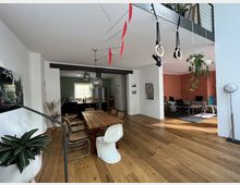 Offener Wohnbereich mit Holzboden, einer Sitzecke mit oranger Wand im Hintergrund und einer großen Holz-Esstafel in der Mitte, umgeben von Stühlen. Oben hängt ein schwarzes Metallgeländer mit Turnringen und Pflanzendekorationen, die von der Decke herabhängen. Im hinteren Bereich ist eine Küche mit grün gestrichener Wand sichtbar.