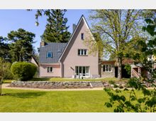 Einfamilienhaus mit steilem Giebel und dunklem Ziegeldach, davor eine kleine Terrasse und eine niedrige Steinmauer mit Stufen. Das Haus steht in einem großen Garten mit Rasenfläche, umgeben von hohen Bäumen und Sträuchern.