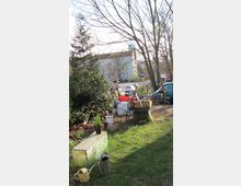 Grüne Gartenszene mit Rasenfläche, einem Metallkasten, Gießkannen und verschiedenen Pflanzen in Töpfen auf einem Tisch. Im Hintergrund stehen mehrere Bäume, ein mehrstöckiges Wohngebäude und parkende Autos an einer Straße.