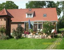 Ein Einfamilienhaus mit rotem Ziegeldach und einer großen Fensterfront, die zum Wintergarten führt. Im Vordergrund befindet sich ein gepflegter Garten mit Blumen, Pflanzen und einer Terrasse, die von Steinbeeten umgeben ist; links steht ein kleines Backsteingebäude mit einem Wetterhahn.