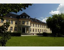 Gelbes Herrenhaus mit symmetrischer Fassade und einem runden Mittelteil, der von Säulen und einem kleinen Eingangsvordach geprägt ist, umgeben von einer gepflegten Rasenfläche und vereinzelten Bäumen unter klarem Himmel.