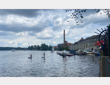Blick auf ein Hafenareal mit anlegenden Motorbooten und Segelbooten, einer historischen, hellen Backsteinfassade mit hohen Schornsteinen im Hintergrund und mehreren Personen, die auf Paddleboards auf dem Wasser stehen. Der Himmel ist bewölkt, und am linken Bildrand ist ein großes Schiff zu sehen.