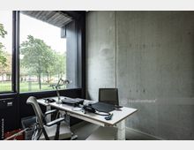 Modernes Büro mit Betonwand, großem Fenster und Blick auf eine grüne Parklandschaft. Im Vordergrund steht ein Schreibtisch mit Bürostuhl, Schreibtischlampe und Arbeitsmaterialien.