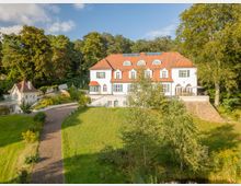 Großes, weißes Herrenhaus mit roten Dachziegeln und grünen Fensterläden, umgeben von gepflegten Rasenflächen und Bäumen. Ein gepflasterter Weg führt durch das parkartige Gelände, das von einem Wald im Hintergrund begrenzt wird.