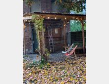 Ein kleiner Außenbereich mit einem hölzernen Pavillon, dekoriert mit Lichterketten, vor der Fassade eines Backsteingebäudes. Darunter befinden sich zwei Liegestühle auf einem mit Herbstlaub bedeckten Boden, umgeben von Pflanzen.