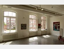 Innenraum einer Galerie mit weiß gestrichenen Wänden, großen Rundbogenfenstern und sichtbaren Heizkörpern. Die Wände sind mit modernem Kunstwerk dekoriert, durch die Fenster ist ein Blick auf rote Ziegelgebäude im Hintergrund zu sehen.