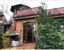 Backsteingebäude mit spitzem Turmdach und dekorativen Holz- und Schmiedeeisenelementen, umgeben von Kletterpflanzen und einem kleinen Baum im Vordergrund. Ein Balkon mit roten, durchbrochen gestalteten Paneelen und ein hölzerner Terrassenboden sind sichtbar.