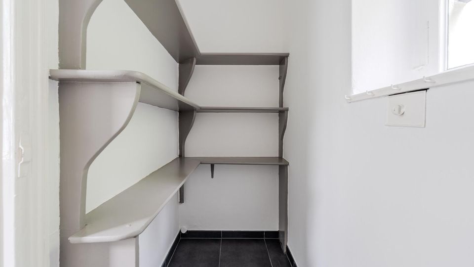 Kleiner Abstellraum mit weißen Wänden und dunkel gefliestem Boden, ausgestattet mit eingebauten Wandregalen in Grau. Rechts befindet sich ein Fenster, das den schmalen Raum mit Tageslicht erhellt.