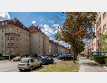 Straßenszene in einer Wohngegend mit mehrstöckigen Wohnhäusern aus hellem Putz und roten Ziegeldächern auf der linken Seite. Rechts sind weitere Wohngebäude mit Balkonen zu sehen, daneben eine Gehwegfläche mit Bäumen und geparkten Autos entlang der Straße.