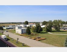 Eine weitläufige Landschaft mit Bürogebäuden und kleinen Gewerbehallen, umgeben von Grünflächen, Bäumen und angelegten Wegen. Im Vordergrund sind ein gepflasterter Weg, ein Parkplatz und ein kleiner Rastplatz mit Sitzbänken zu sehen.