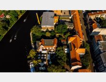 Luftaufnahme eines Stadtviertels mit einem Fluss auf der linken Seite, auf dem mehrere Boote fahren. Rechts vom Fluss befinden sich Gebäude mit roten Ziegeldächern, eine grün bewachsene Parkanlage und ein Bereich mit weißen Sonnenschirmen, der auf ein Café oder Restaurant hindeutet.
