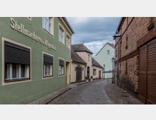 Schmale Kopfsteinpflasterstraße in einem historischen Wohnviertel mit niedrigen Häusern. Links ein grünes Gebäude mit der Aufschrift „Stellmacherei u. Wagenbau“, rechts eine Ziegelfassade; im Hintergrund moderne und ältere Gebäude sichtbar.
