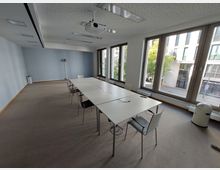 Konferenzraum mit großem rechteckigem Tisch in der Mitte, umgeben von Stühlen. Der Raum hat eine blaue Wand, helle Decke mit integrierter Beleuchtung, grauen Teppichboden und große Fenster, die Tageslicht hereinlassen und Blick auf eine Straße und umliegende Gebäude bieten.