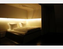 Ein modern eingerichtetes Schlafzimmer mit einem schwebenden Doppelbett, beleuchtet durch indirekte, warmweiße LED-Leuchten an der Wand. Der Raum ist durch dunkle Böden und Vorhänge schlicht gestaltet.
