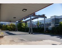 Großer asphaltierter Platz mit Überdachung und Metallkonstruktionen, umgeben von Industriegebäuden mit großen Glasfenstern. Im Hintergrund sind Bäume und Büsche zu sehen, die teilweise die Fassaden der Gebäude bewachsen.