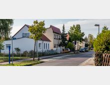 Straßenszene in einem Wohngebiet mit Einfamilienhäusern und Vorgärten, die von Zäunen umgeben sind. Die Straße ist asphaltiert und von geparkten Autos sowie mehreren Bäumen gesäumt, während Gehwege auf beiden Seiten sichtbar sind.