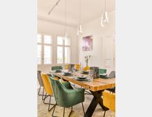 Esszimmer mit modernem Design, ausgestattet mit einem großen Tisch aus Holz und gepolsterten Stühlen in Grün und Gelb. Die weißen Wände sind mit dekorativen Elementen wie einem bunten Wandbild und hängenden Glasleuchten verziert, während große Fenster viel Tageslicht einlassen.