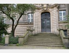 Historisches Gebäude mit steinerner Treppe, die zu einem großen, verzierten Schmiedeeisentor führt. Die Fassade ist aus hellem Stein und wird von Fenstern mit weißen Rahmen sowie dekorativen Elementen geprägt, umgeben von Bäumen und einem schmiedeeisernen Zaun.