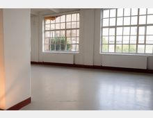 Leerer, heller Innenraum mit weiß gestrichenen Wänden und glattem Boden, geprägt von großen Sprossenfenstern. Unter den Fenstern verlaufen Heizkörper und rote Sockelleisten, draußen sind Gebäude und etwas Grün zu sehen.