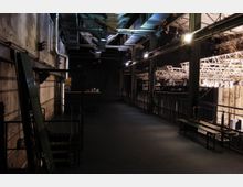 Industrieartige Backstage-Bühnenumgebung mit sichtbaren Metallkonstruktionen, Lüftungsrohren und Beleuchtungsträgern. Rechts befindet sich ein erhöhter Bereich mit Geländern, unter dem ein Bühnenaufbau mit zahlreichen Scheinwerfern erkennbar ist.