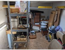 Ein kleiner, vollgestellter Abstellraum mit Metallregalen, auf denen Kartons, Werkzeuge und Behälter stehen. Im Hintergrund liegen gestapelte Gegenstände, darunter Autoteile, Kartons und Möbelstücke; an den Seiten lehnen Kartonplatten an der Wand.