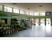 Innenraum einer hellen Bahnhofshalle mit großen Fenstern unter der Decke und grünen Kacheln an den Wänden. Im Vordergrund befinden sich ein Blumenladen mit bunten Pflanzen und Verkaufsständer mit Zeitschriften, während durch die geöffneten Türen ein Blick auf eine belebte Außenumgebung sichtbar wird.