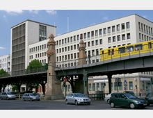 Straßenszene in einer Stadt mit erhöht verlaufender U-Bahnstrecke, auf der eine gelbe U-Bahn fährt. Im Vordergrund ist eine befahrene Straßenkreuzung zu sehen, flankiert von Steinsäulen mit Verzierungen, und im Hintergrund befindet sich ein mehrstöckiges Bürogebäude mit einfacher, rechteckiger Fassade.