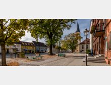 Kleiner Platz in einer historischen Altstadt mit Kopfsteinpflaster, großen Bäumen, bunten Häuserfassaden und einer Kirche mit spitzem Turm im Hintergrund. Im Vordergrund stehen Sitzbänke und Tische, daneben Straßenlaternen und Blumen an einem Backsteingebäude.