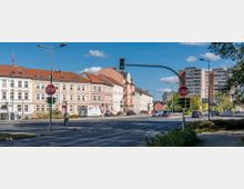 Straßenkreuzung in einer Stadt mit mehrspurigen Fahrbahnen, Ampeln und Stoppschildern. Im Hintergrund befinden sich historische Wohnhäuser mit Ziegeldächern sowie moderne Wohnblöcke, umgeben von Bäumen und Grünflächen.