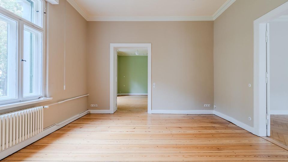 Leerer Wohnraum mit hellem Holzfußboden, hohen Fenstern an der linken Wand und einem Heizkörper darunter. In der Mitte führt eine Türöffnung in ein weiteres Zimmer mit grün gestrichener Wand; weiße Türrahmen und Stuckleisten an der Decke sind sichtbar.