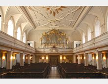 Innenraum einer historischen Kirche mit einer prunkvollen, goldverzierten Orgel auf der Empore im Hintergrund. Hohe Rundbögen, ein reich dekoriertes Deckenrelief und Holzbankreihen prägen den Raum.