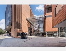 Moderne Architektur am Potsdamer Platz in Berlin mit roten Fassaden, verglastem Eingangsbereich und dem Schriftzug "Potsdamer Platz". Im Vordergrund Gehweg, ein futuristischer Pavillon und eine Glasüberdachung vor dem Eingang.