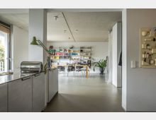 Offener Wohnraum mit minimalistischem Design, bestehend aus einer Küche mit Metallarbeitsfläche und angrenzendem Essbereich mit einem großen Holztisch und Bücherregalen an der Wand. Auf dem Boden steht eine große Zimmerpflanze, und das Licht fällt durch ein bodentiefes Fenster auf den Betonboden.