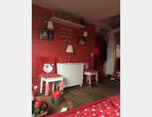 Ein kleines Zimmer mit weihnachtlicher Dekoration in Rot und Weiß. Die Wände sind mit rot-weiß gepunkteter Tapete bedeckt, Stühle haben Überzüge mit Weihnachtsmotiven, und auf einem Regal stehen Gläser und weihnachtliche Dekorartikel.