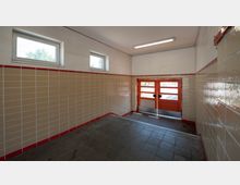 Innenraum einer kleinen Garage oder eines Abstellraums mit hellbeigen Fliesenwänden und roten Fliesenkanten, einem grauen Boden und einem orangefarbenen, verglasten Tor am Ende. Zwei rechteckige Fenster an der oberen Wand lassen Tageslicht herein.