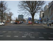 Straßenansicht mit einem Parkplatz im Hintergrund, umgeben von mehreren Bäumen, modernen Gebäuden und Wandgraffiti. Im Vordergrund befindet sich eine Straße mit Fahrbahnmarkierungen für Autos und Fahrräder.