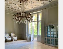 Eleganter Innenraum mit einem großen Kristalllüster, einer verzierten weißen Holzdecke und großen Fenstern, die einen Blick ins Grüne ermöglichen. Im Raum stehen gepolsterte Sessel unter Plastikabdeckung sowie eine weiße Vitrine mit Glasfronten.