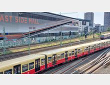 Blick auf eine Bahnstrecke mit einem vorbeifahrenden gelb-roten S-Bahn-Zug der Deutschen Bahn. Im Hintergrund steht ein modernes Gebäude mit der Aufschrift „East Side Mall“, daneben befinden sich weitere Bauwerke und Baukräne.