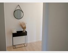 Schlichter Flur mit hellem Holzfußboden und weißen Wänden, dekoriert mit einem runden Wandspiegel und einem minimalistischen schwarzen Beistelltisch. Auf dem Tisch stehen eine Vase mit Trockenblumen und einige Zeitschriften.