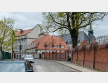 Straßenszene mit Kopfsteinpflaster, flankiert von einer roten Backsteinmauer und Gebäuden mit roten Ziegeldächern und verzierten Giebeln; im Hintergrund ein Kirchturm und Bäume mit frischem Laub.