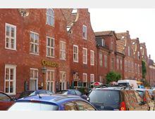 Straßenszene mit historischer roter Backsteinarchitektur und mehreren Gebäuden mit dekorativen Giebeln. Im Vordergrund geparkte Autos, darunter Schilder von Geschäften oder Restaurants wie "Charlotte".