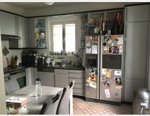 Moderne Küche mit grauen Schränken und einer Arbeitsfläche aus schwarzem Material. Im Vordergrund ein Esstisch mit vier grauen Stühlen, im Hintergrund ein großer Kühlschrank mit Magneten und eine Spüle unter einem Fenster.