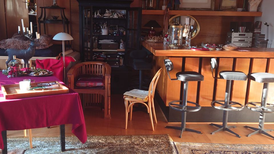 Ein Wohnzimmer oder gemütlicher Aufenthaltsraum mit Holzelementen, einer kleinen Bar mit Barhockern, bunten Teppichen auf Holzboden und dekorativ platzierten Möbeln, einschließlich eines Regals mit Figuren und einer Tischfläche mit roten Tischdecken.