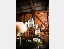 Ein rustikaler Innenraum mit Holzwänden und sichtbaren Dachbalken, dekoriert mit Wassergefäßen aus Glas auf Holzkisten. Im Vordergrund sind Blätter und Blumenarrangements zu erkennen, während im Hintergrund landwirtschaftliche Werkzeuge und ein Wandregal sichtbar sind.