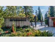 Eingang zu einem Friedhof mit einer Steinmauer und einem offenen Metalltor mit geschwungenem Design. Im Vordergrund sind grüne und rote Pflanzen zu sehen, im Hintergrund hohe Bäume und ein gepflasterter Weg.