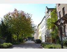 Ruhige Wohnstraße mit Kopfsteinpflaster, gesäumt von Gebäuden mit historischen Fassaden auf der rechten Seite und großen Bäumen auf der linken Seite. Am Straßenrand sind parkende Autos und gepflegte Grünflächen zu sehen.