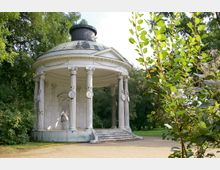 Klassizistischer Pavillon mit einer runden Kuppel und korinthischen Säulen, umgeben von Bäumen und Büschen in einem Park. Im Inneren des Pavillons befindet sich eine Statue auf einem Sockel, daneben führt eine kleine Treppe hinauf.